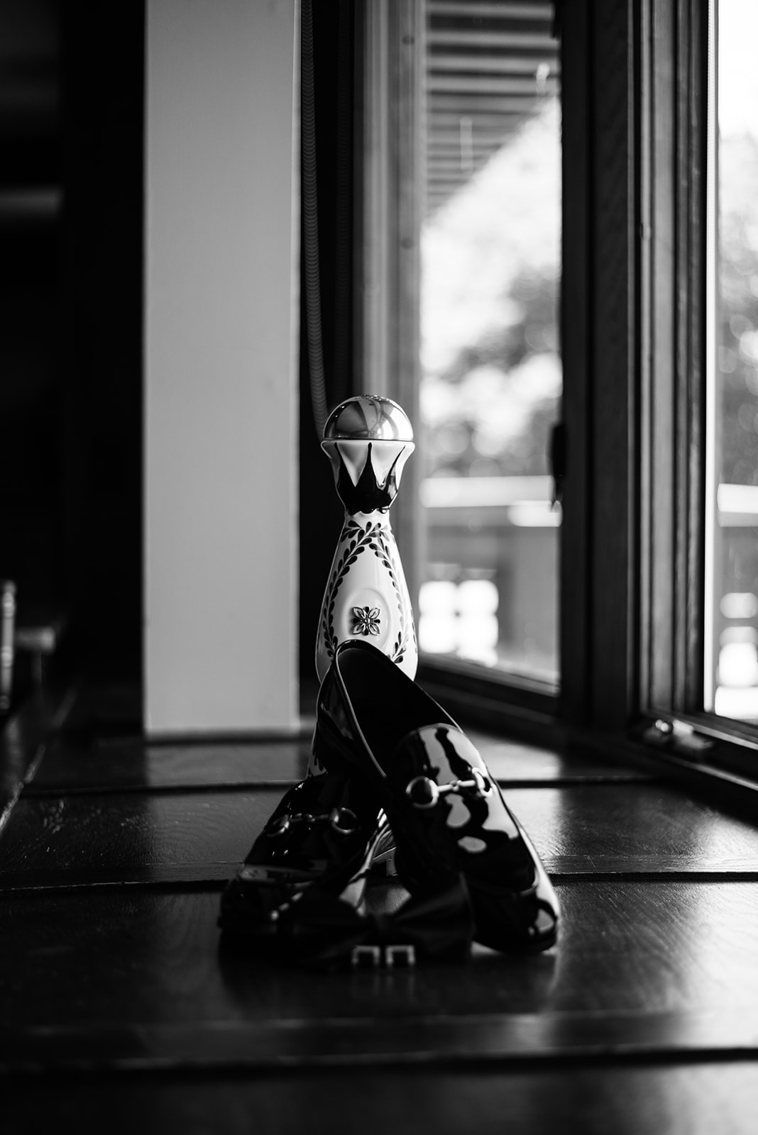 Black and white film photo of the groom's patent loafers, bow tie, and a tequila bottle on a windowsill.